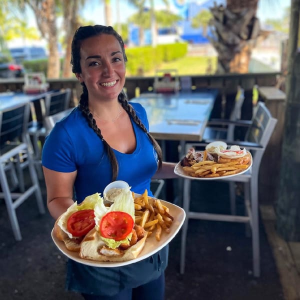 Smiling server at Mangia Clam Bar holding two plates of seafood sandwiches and fries on an outdoor patio.