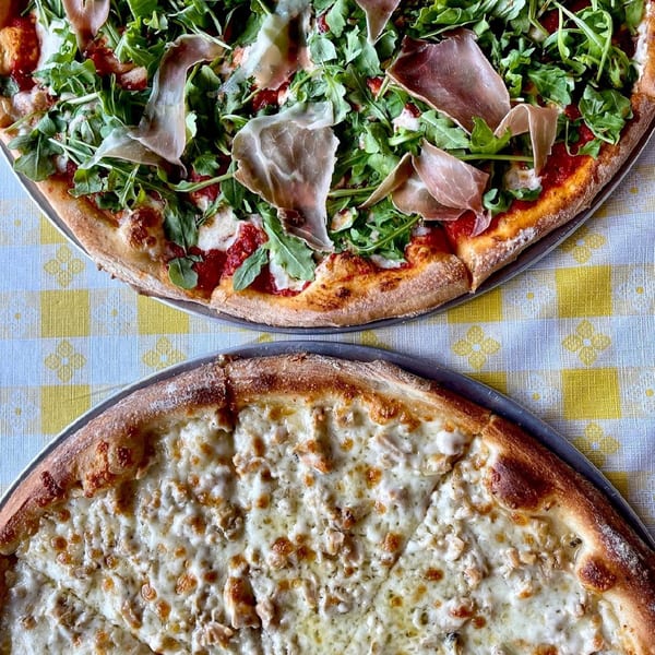 Two pizzas on a checkered tablecloth: one with prosciutto and arugula, the other a white clam pizza.
