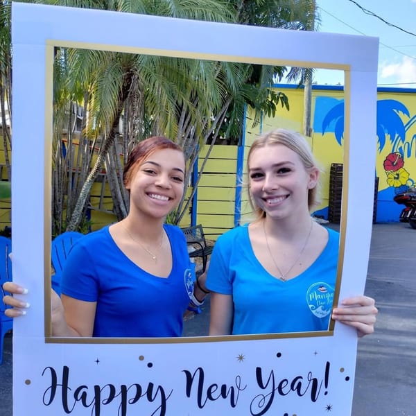 Two smiling women in blue Mangia Clam Bar shirts hold a "Happy New Year!" frame outdoors.