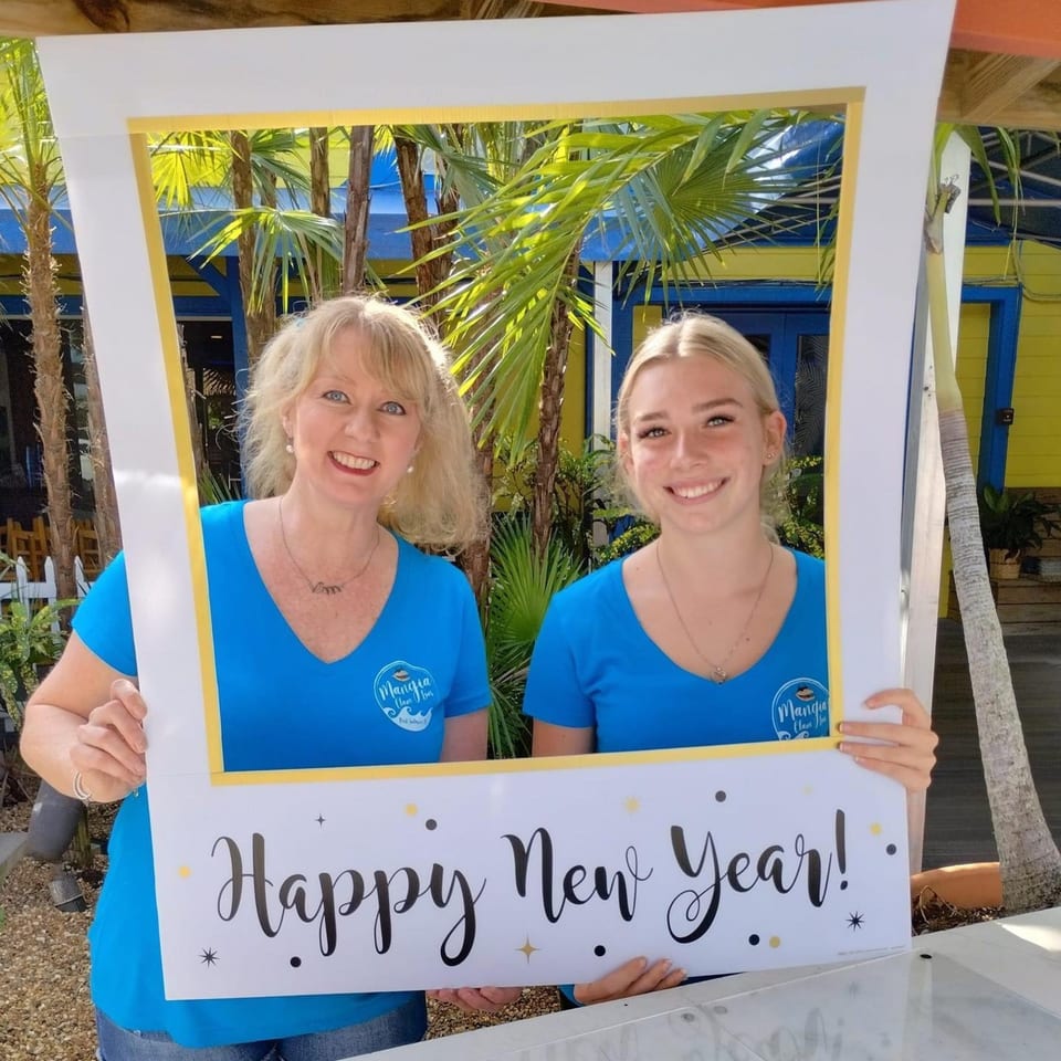 Two smiling women in blue Mangia Clam Bar shirts hold a "Happy New Year!" frame outdoors.
