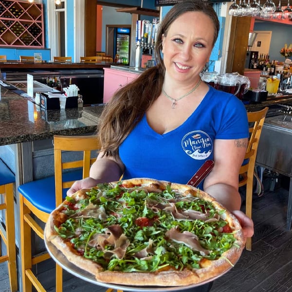 A smiling Mangia Clam Bar server holding a large pizza topped with prosciutto and arugula.