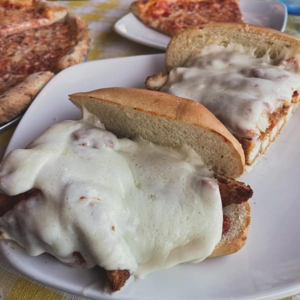 Two Mangia Clam Bar chicken parmesan sandwiches with melted cheese, and pizza in the background.