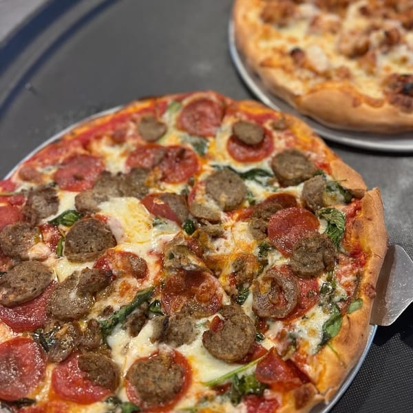 Two pizzas, one with pepperoni, sausage, and spinach, and another with chicken, on dark trays.