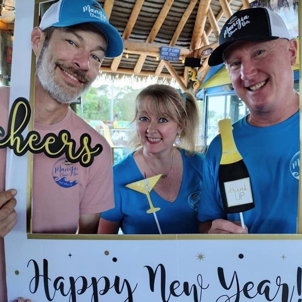 Three smiling Mangia Clam Bar staff members pose in a "Happy New Year" frame with drink props.